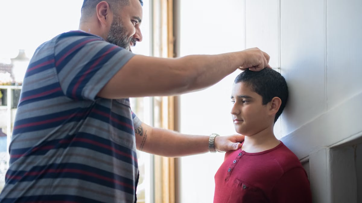 A father measuring his son's height against a wall in their home, capturing a family growth moment.